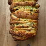 A cheesy, herb-filled pull-apart loaf on a wooden cutting board, golden and bubbling.