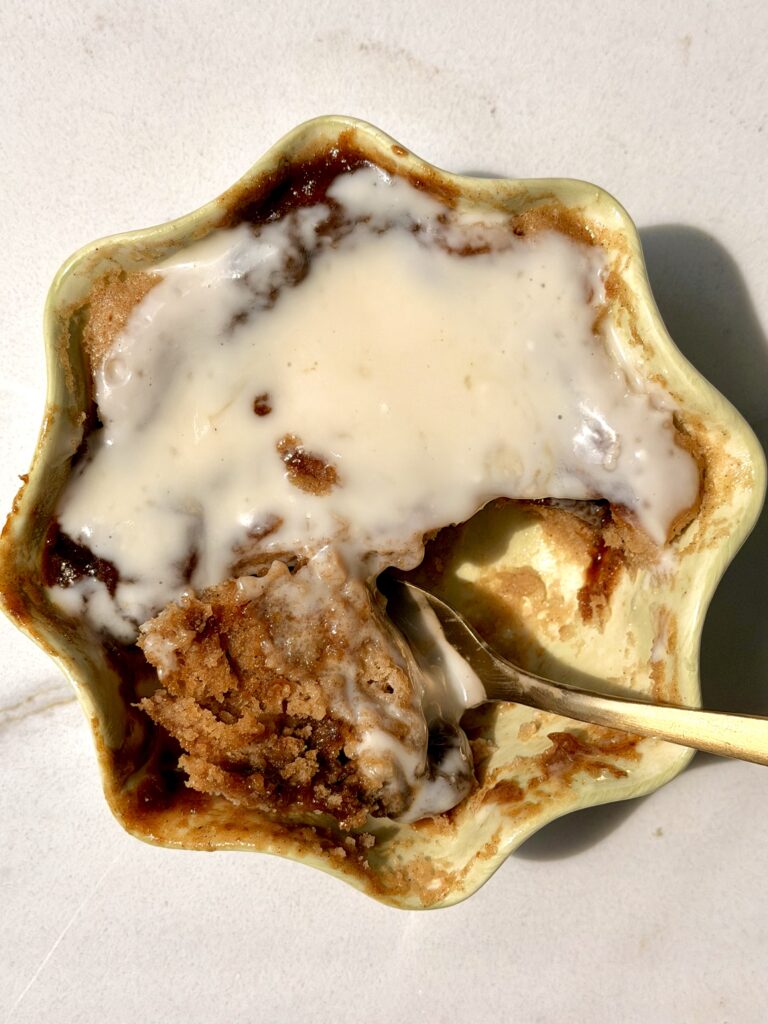High protein cinnamon roll mug cake in a sage green ramekin topped with creamy vanilla glaze, with a gold spoon scooping into the soft cinnamon center, shot from overhead on a white marble surface.
